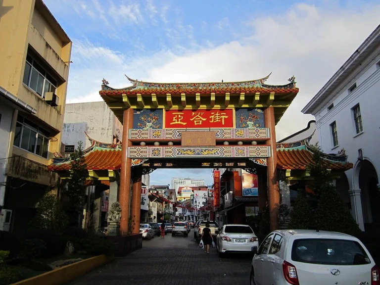 A large, ornate red and gold Chinese gateway over a city street in Kuching with cars parked along the sides of the narrow urban street.